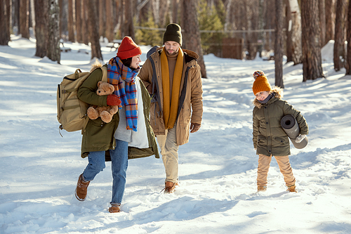 Mother and little daughter in warm winterwear having fun in snowdrift on their way to country house, father following them on winter day