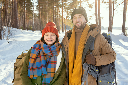 Young smiling couple in warm winterwear standing in front of camera against snow and mixed forest and looking at you on sunny day