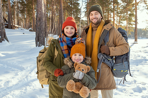 Cheerful young family of three in warm winterwear standing in front of camera against natural environment and looking at you outside