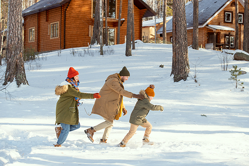 Side view of cheerful young man, his wife and their cute little daughter in warm winterwear having fun in snowdrift on their way to country house