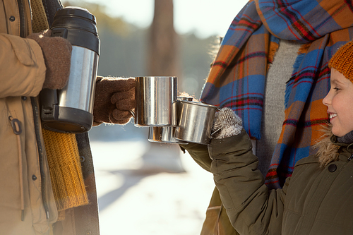 Happy little girl and her parents in warm winterwear clinking with metallic mugs with hot tea while getting warm outside on frosty winter day