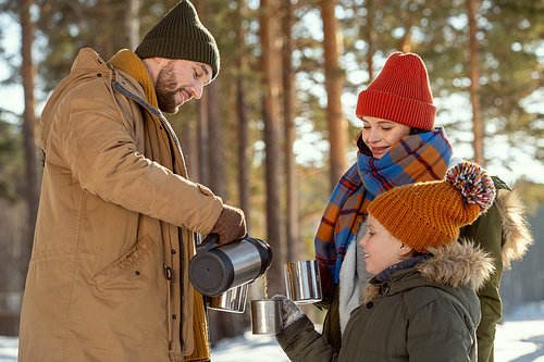 Happy young man pouring hot tea into metallic mugs of his wife and daughter while standing in front of camera against pine trees