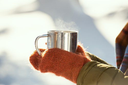 Hands of young female in warm winterwear holding metallic mug with hot tea while standing in front of camera against shadows on snow