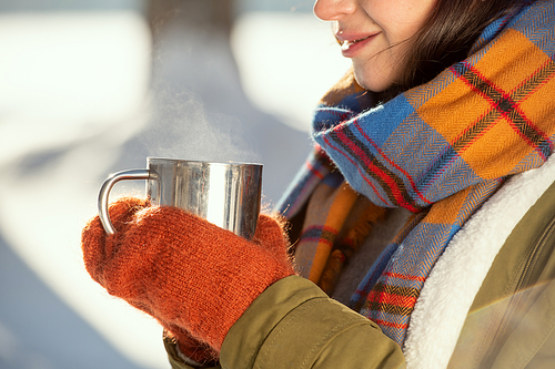 Hands of young female in warm jacket, scarf and woolen knitted mittens holding metallic mug with hot tea while standing in front of camera