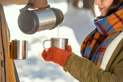 Young female in warm winterwear holding metallic mug while her husband pouring hot tea from thermos in front of camera against snow