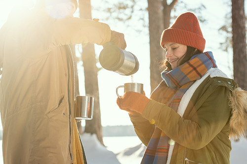 Happy young female in warm winterwear holding metallic mug while her husband pouring hot tea in front of camera against pine trees