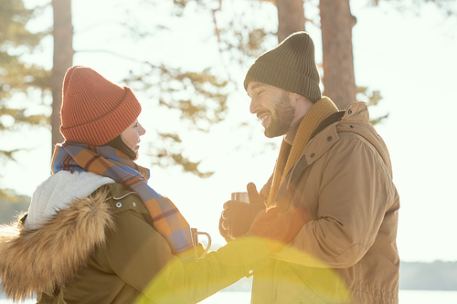 Happy young affectionate couple in warm winterwear having hot tea while standing in front of one another, talking and enjoying winter weekend