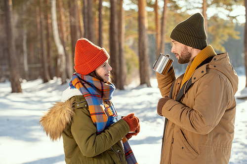 Side view of young couple in warm winterwear having hot tea while standing in front of one another, talking and enjoying winter weekend