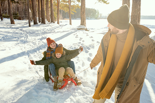 Happy young father in warm winterwear pulling sledge with his laughing wife and their cute little daughter while having fun on winter day