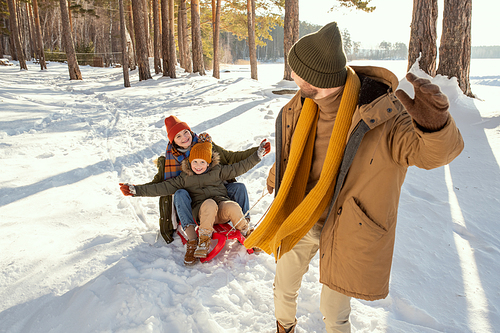 Cheerful young man in warm winterwear pulling sledge with his laughing wife and their cute little daughter while having fun on winter day