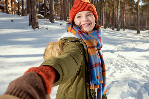 Happy young woman in warm winterwear holding her husband by hand and looking at him with toothy smile while inviting to follow her