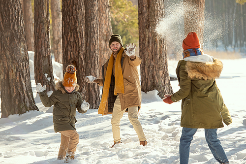 Cheerful young man, his wife and their cute little daughter in warm winterwear playing snowballs while having fun in snowdrift