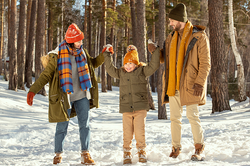 Happy young parents in warm winterwear holding their cute smiling little daughter by hands while helping her to jump in snowdrift