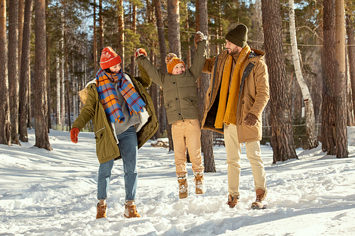 Happy young parents in warm winterwear holding their jumping little daughter by hands while having fun in snowdrift among pines