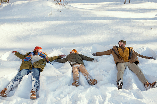 Cheerful young man, his wife and their cute little daughter in warm winterwear lying on snow while having fun in snowdrift on winter day