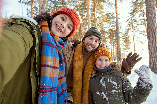 Happy young parents and their cute little daughter in warm winterwear making selfie against natural environment on frosty winter day