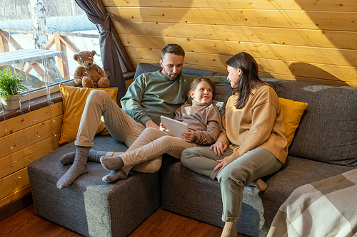 Happy young family of father, mother and little daughter in casualwear sitting on sofa inside country house and watching online movie