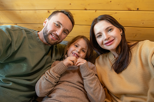 Happy young family of father, mother and little daughter in casualwear sitting on sofa inside country house and looking at you with smiles