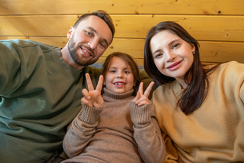 Cheerful father, mother and little daughter in casualwear relaxing in front of camera inside country house and looking at you with smiles