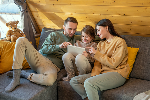 Happy young family of three in casualwear sitting on sofa inside country house and watching online movie while pointing at tablet screen