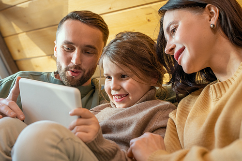 Happy young family of three in casualwear relaxing inside their country house and watching online movie while looking at tablet screen