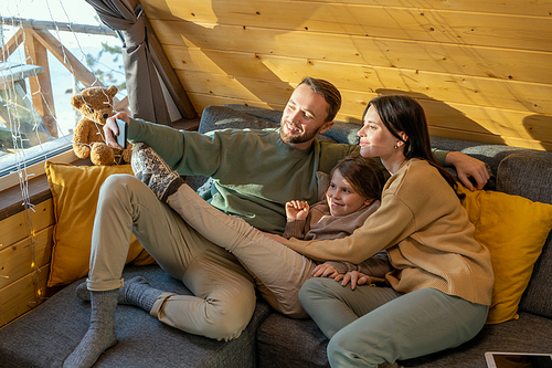 Happy young parents and their cute little daughter in casualwear making selfie while relaxing on large comfortable sofa in country house