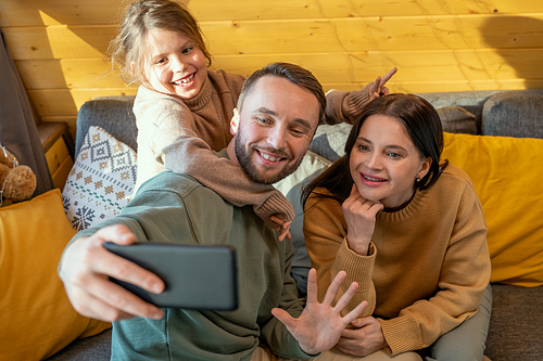 Young cheerful couple and cute little daughter in casualwear making selfie or communicating through video chat in their country house