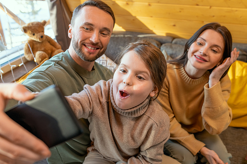 Funny and cute little girl and her cheerful parents in casualwear making selfie or communicating through video chat in their country house