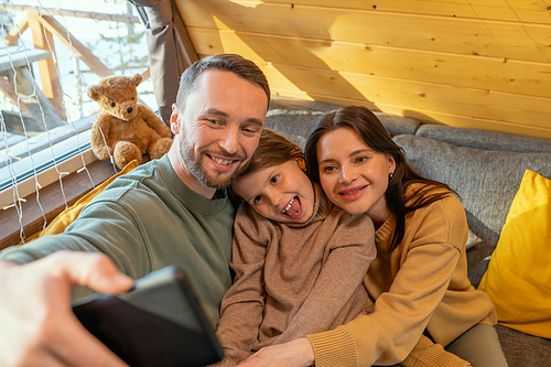 Funny little girl and her cheerful parents making selfie or communicating through video chat while relaxing and having fun in their country house