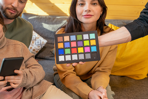 Hand of young man holding color palette in front of young brunette woman, her husband and daughter relaxing in country house