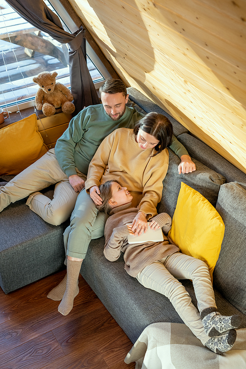 Happy young relaxed family of father, mother and little daughter in casualwear lying on large sofa inside their country house