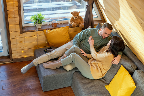 Happy young relaxed husband and wife in casualwear lying on large sofa inside their country house, enjoying weekend and having chat