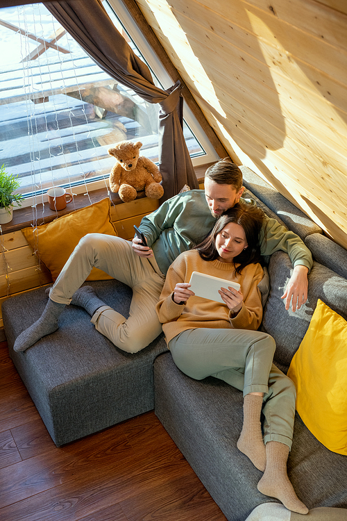 Happy young relaxed husband and wife in casualwear lying on large sofa inside their country house, enjoying rest and watching online movie