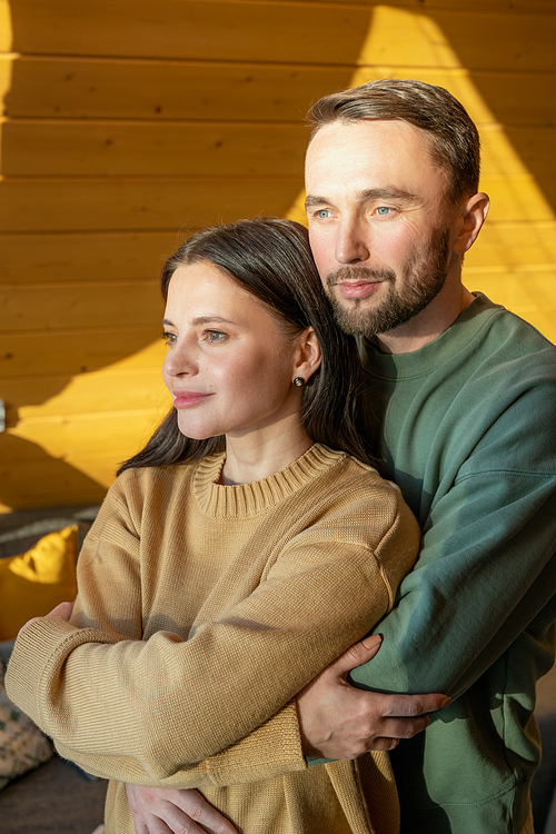 Young affectionate husband embracing wife while standing in front of large window and enjoying weekend in their country house