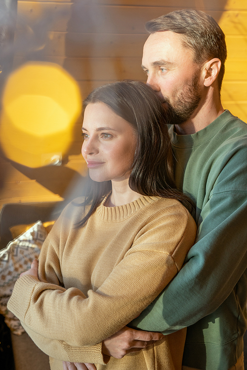 Young affectionate man embracing his wife while standing behind her in front of large window and enjoying weekend in their country house