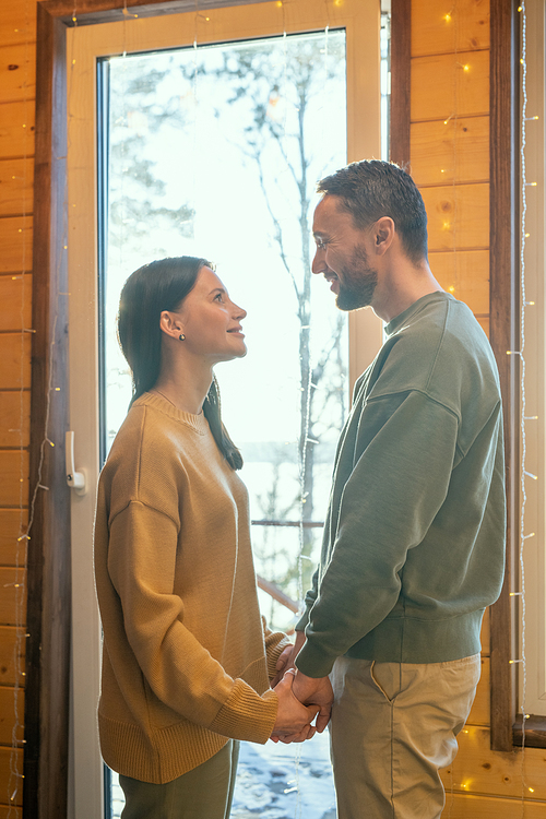 Happy young affectionate husband and wife holding by hands while standing in front of large window inside their country house