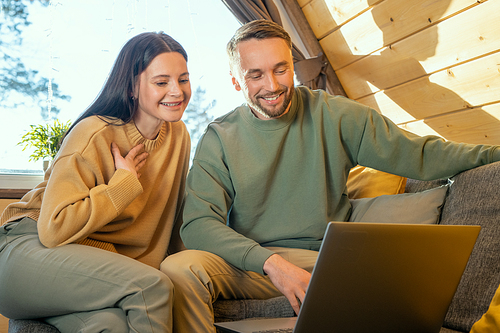 Happy young affectionate couple in casualwear sitting on large sofa in front of laptop display and communicating through video chat