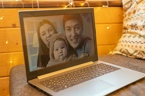 Happy young family of three on display of laptop standing on large soft comfortable sofa inside country house during online communication