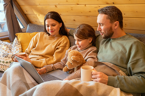 Happy young family in casualwear sitting on sofa under checkered plaid inside country house and watching online movie on laptop display