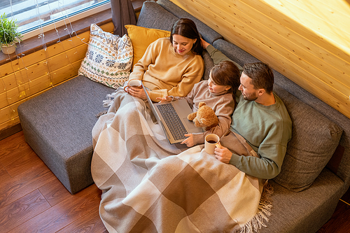 Happy young family in sweaters relaxing on large comfortable sofa under checkered plaid inside country house and watching online movie