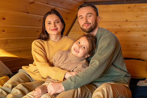 Happy young affectionate father, mother and little daughter in casualwear sitting on sofa inside country house and looking at you with smiles