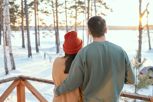 Rear view of young affectionate man in sweater embracing his wife while both standing in front of camera on patio of their country house