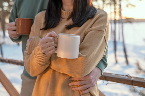 Young affectionate man embracing his wife with mug of hot tea while both standing in front of camera on patio of their country house