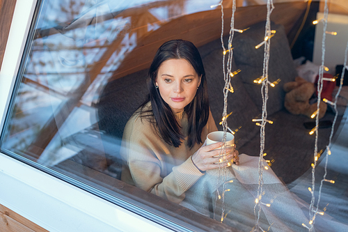 Young serene brunette female in sweater having hot tea while sitting on couch by large window of country house on winter weekend