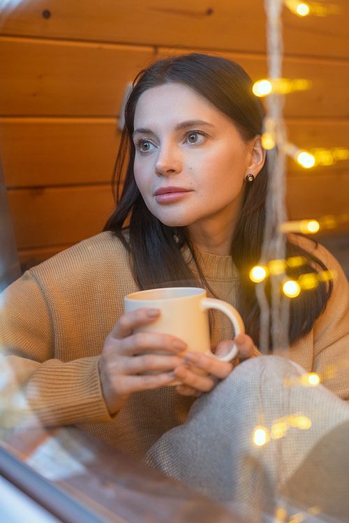 Young serene brunette female in sweater having hot tea while sitting by large window of country house and enjoying winter weekend