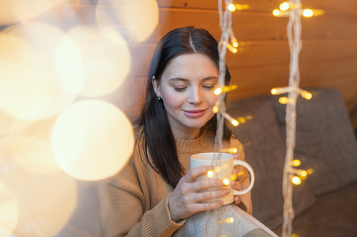 Young serene brunette woman with mug of hot tea sitting on couch by large window of country house on winter weekend and enjoying rest