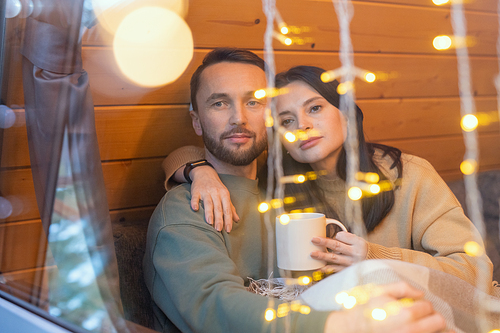 Young serene affectionate couple in sweaters sitting on couch by large window of country house on winter weekend and enjoying rest