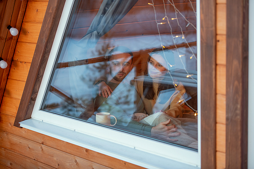 Young serene affectionate couple in sweaters looking through large window of country house on winter weekend while enjoying rest