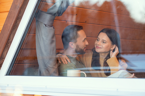 Young serene affectionate couple sitting by large window of country house on winter weekend, looking at one another and interacting