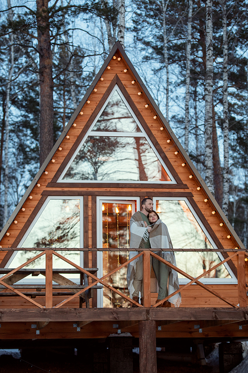 Young affectionate couple wrapped in plaid standing by new large country house on background of pines and birches and looking at sunrise